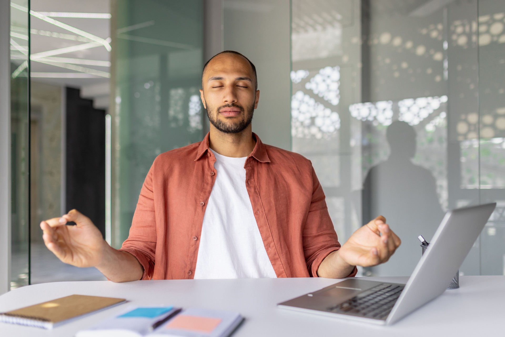 Man practicing mindfulness meditation in modern office setting with laptop and notebooks. Meditation promotes stress relief and focus during work. Achieve inner peace and relaxation at work.